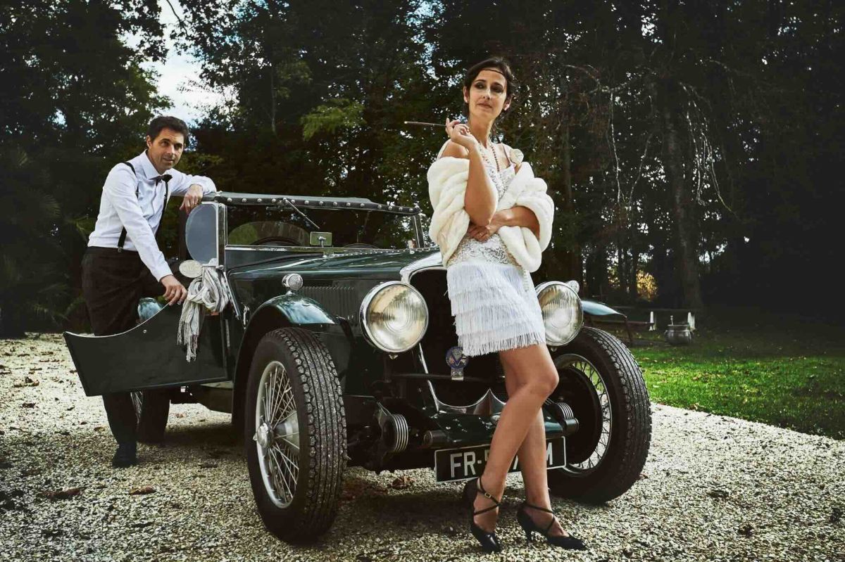 Photographie d'exposition 2025 : couple en tenue "Années Folles" posant devant une voiture ancienne verte, ambiance Gatsby ou film d'époque.