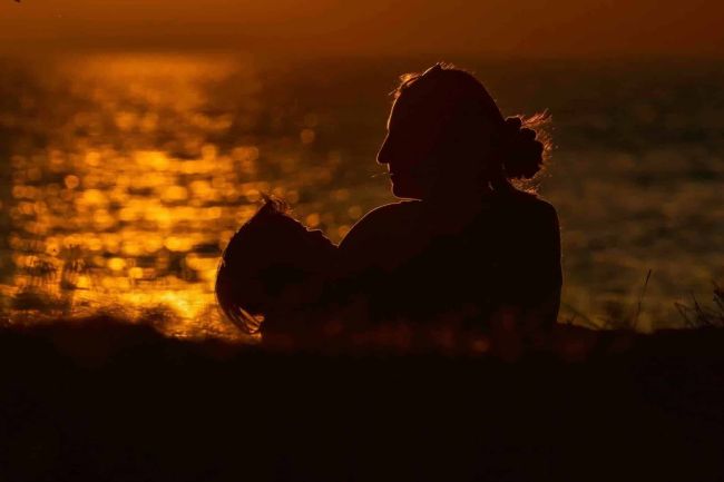 Photographie exposition 2024 Fragilité : silhouettes d'une mère et de son enfant au coucher de soleil sur l'eau, tendresse et lien fragile.