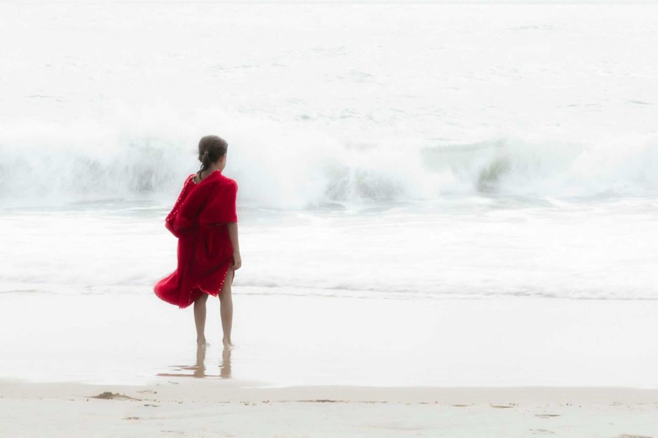 Photographie exposition 2024 Fragilité : femme en robe rouge face à une grande vague blanche et mouvante, force de la mer.